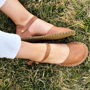 May include: A pair of tan leather sandals featuring a rounded toe, a thin ankle strap with a small metal buckle, and visible light-colored stitching along the edge of the sole. The sandals have a flat design and a smooth, matte finish.