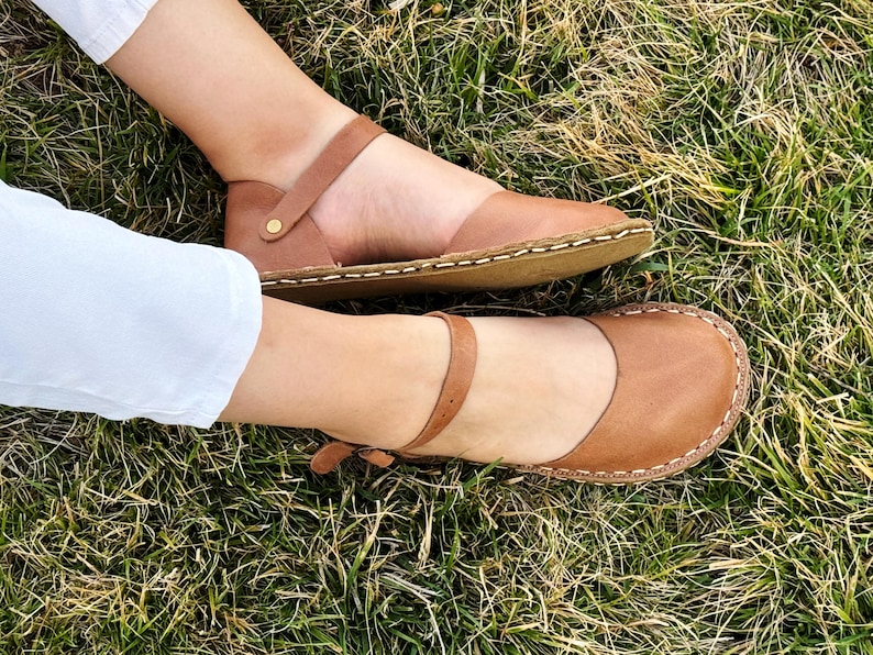 May include: A pair of tan leather sandals featuring a rounded toe, a thin ankle strap with a small metal buckle, and visible light-colored stitching along the edge of the sole. The sandals have a flat design and a smooth, matte finish.