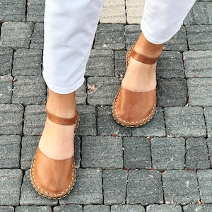 May include: A pair of tan leather sandals featuring a rounded closed-toe design, a thin ankle strap with a buckle closure, and visible light-colored stitching along the sole edge, worn with white trousers on a gray stone paved surface.