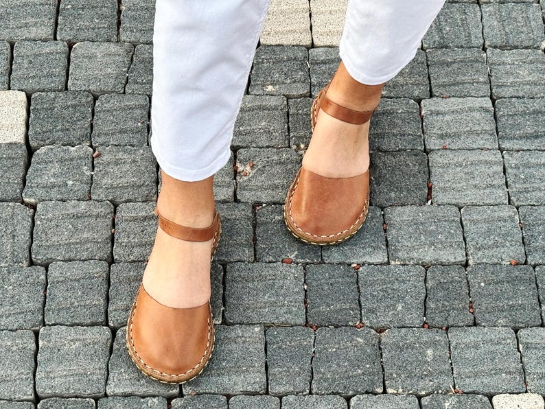 May include: A pair of tan leather sandals featuring a rounded closed-toe design, a thin ankle strap with a buckle closure, and visible light-colored stitching along the sole edge, worn with white trousers on a gray stone paved surface.