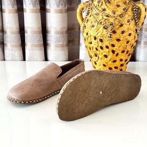 May include: A pair of light brown slip-on shoes with a smooth upper and a textured, flexible sole. One shoe is positioned to show the top, while the other is placed on its side to reveal the bottom surface, featuring visible stitching along the edge.