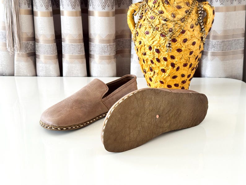 May include: A pair of light brown slip-on shoes with a smooth upper and a textured, flexible sole. One shoe is positioned to show the top, while the other is placed on its side to reveal the bottom surface, featuring visible stitching along the edge.
