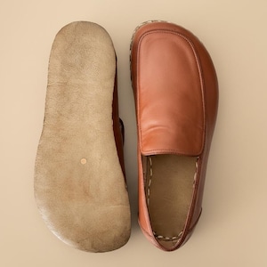 May include: A pair of tan leather slip-on shoes shown from two different angles. One shoe displays the smooth, rounded brown leather upper, while the other shoe shows the light-coloured, textured suede sole with visible stitching along the edge.