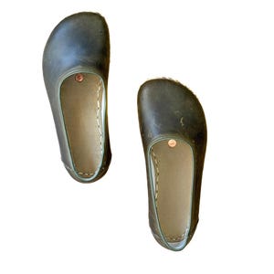 May include: A pair of dark olive green slip-on shoes with a rounded toe box and a wide opening. The interior features a light tan lining with visible white stitching along the edges and a small circular copper-coloured rivet near the opening.