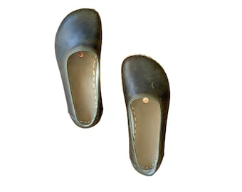 May include: A pair of dark olive green slip-on shoes with a rounded toe box and a wide opening. The interior features a light tan lining with visible white stitching along the edges and a small circular copper-colored rivet near the opening.