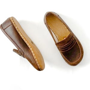 May include: A pair of brown leather loafers featuring a classic penny slot design across the vamp, visible light-colored stitching along the sole edge, and a smooth interior lining, presented as one side view and one top-down view.