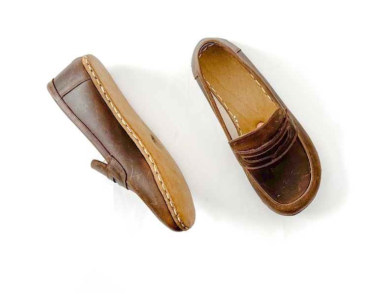 May include: A pair of brown leather loafers featuring a classic penny slot design across the vamp, visible light-colored stitching along the sole edge, and a smooth interior lining, presented as one side view and one top-down view.