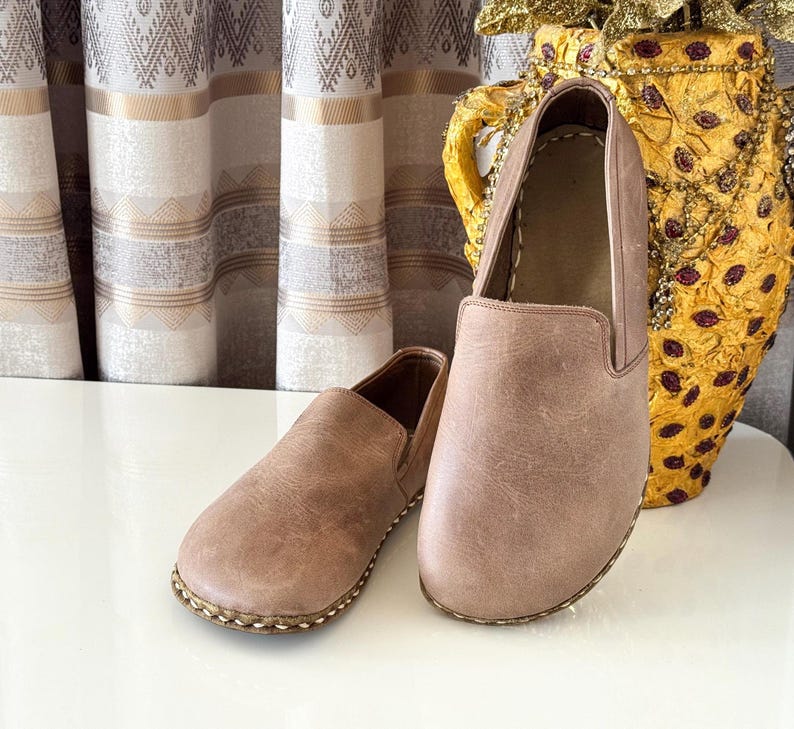 May include: A pair of light brown slip-on shoes featuring a smooth matte finish, rounded toes, and visible thick stitching along the sole edges. The shoes have a simple, low-profile design with elastic side panels for a secure fit and flat soles.