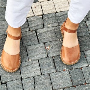 May include: A pair of tan leather sandals featuring a rounded closed-toe design, a thin ankle strap with a buckle closure, and visible light-colored stitching along the sole edge. The sandals have a smooth finish and a flat, flexible construction.