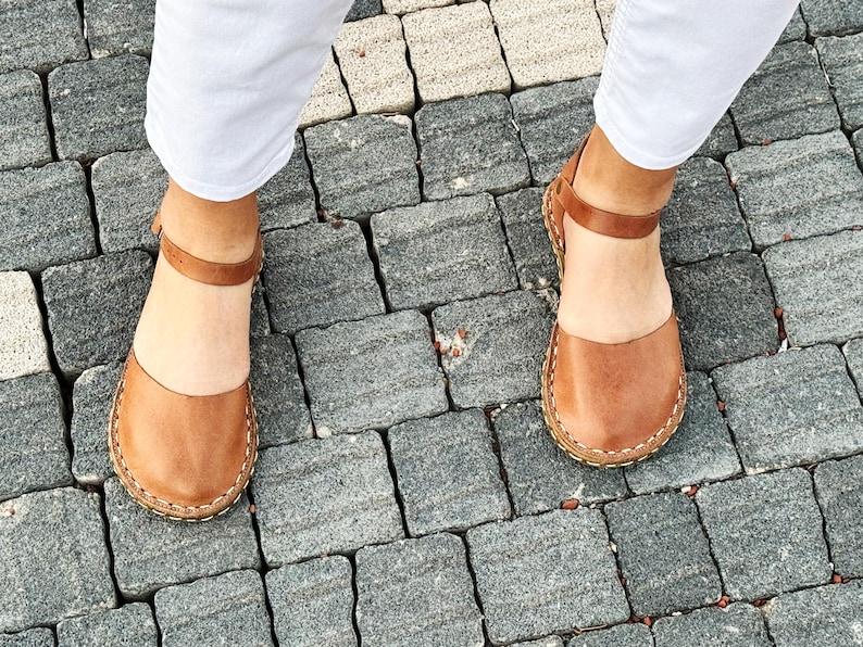 May include: A pair of tan leather sandals featuring a rounded closed-toe design, a thin ankle strap with a buckle closure, and visible light-colored stitching along the sole edge. The sandals have a smooth finish and a flat, flexible construction.