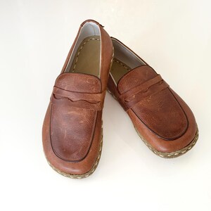 May include: A pair of brown leather loafers featuring a rounded toe, a scalloped strap across the vamp, and visible light-coloured stitching along the sole edge. The interior lining is a light tan colour, and the leather has a natural textured finish.