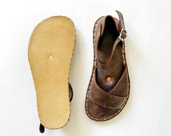 May include: A pair of brown leather sandals featuring a crisscross strap design, an adjustable ankle strap with a silver-tone metal buckle, visible white stitching along the sole edge, and a light tan textured sole on the underside.