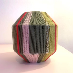 Può includere: Un vaso decorativo e geometrico avvolto in filo verde oliva con strisce verticali rosse, rosa e oro. Il vaso ha un aspetto unico e strutturato ed è adatto per l'arredamento della casa.