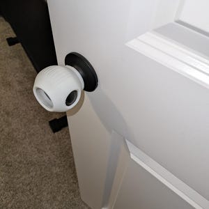 May include: A white door knob safety cover with a rounded shape and two eye-like openings. The cover is attached to a white door with a black door knob. The door is set against a neutral-colored carpet.