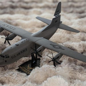 May include: A gray model of a U.S. Air Force C-130 Hercules aircraft. The model has four propellers, wings, and the words "U.S. AIR FORCE" printed on the side. The model is mounted on a black stand with text.