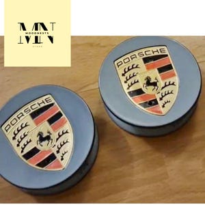 May include: Two light blue Porsche hubcaps with the Porsche emblem in the center. The emblem features a black horse, red and black stripes, and the word "PORSCHE" in black.