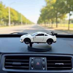 May include: A white toy sports car with black wheels and a rear spoiler is displayed on a dashboard. The car is mounted on a small, dark gray stand. The background shows a road and trees.