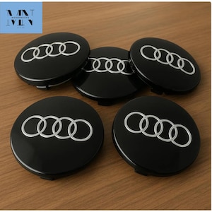 May include: Five black Audi wheel centre caps, each featuring the silver Audi logo of four interlocking rings. The caps are displayed on a wooden surface. The caps are new.