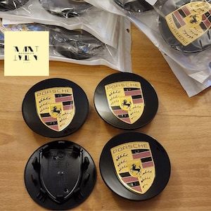 May include: Four black Porsche wheel centre caps featuring the Porsche emblem in gold, red, and black. The caps are circular with a glossy finish. The image also shows a logo that says "MNT MOODNEST STORE".