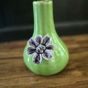 May include: A small, green ceramic vase with a pear-shaped body and a narrow neck. A silver-colored flower is affixed to the front of the vase. The vase is sitting on a wooden surface.