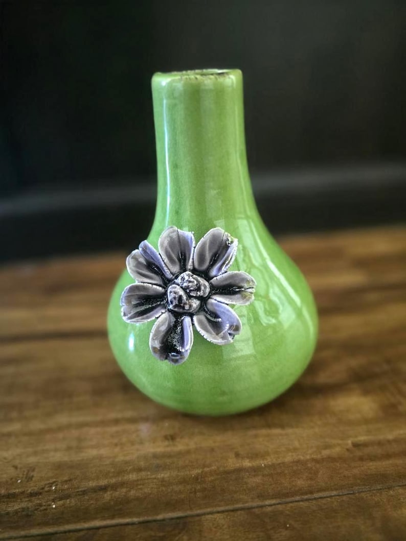 May include: A small, green ceramic vase with a pear-shaped body and a narrow neck. A silver-colored flower is affixed to the front of the vase. The vase is sitting on a wooden surface.
