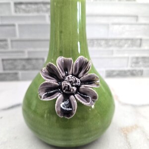 May include: A green ceramic vase with a bulbous base and a slender neck. A detailed, dark purple flower is affixed to the front of the vase. The vase has a glossy finish and is likely for decorative purposes.