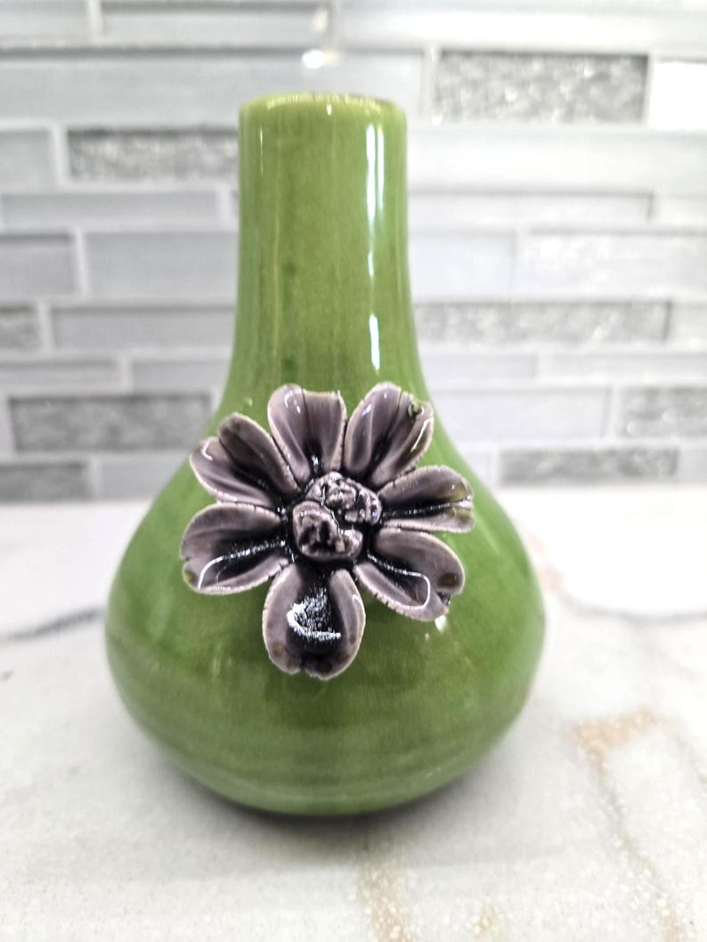 May include: A green ceramic vase with a bulbous base and a slender neck. A detailed, dark purple flower is affixed to the front of the vase. The vase has a glossy finish and is likely for decorative purposes.