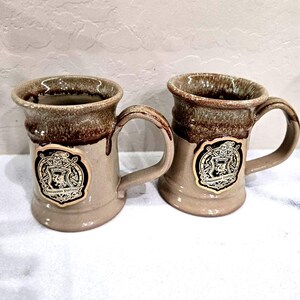 May include: Two ceramic mugs with a tan base and a dark brown drip glaze around the rim. Each mug features a circular emblem with a black background, a gold-colored crest, and the text Arizona Renaissance Festival 2024 printed on the front.