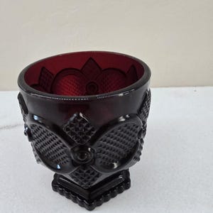 May include: A ruby red glass bowl with a detailed floral and geometric pattern. The bowl has a square base and a decorative design around the sides. The interior of the bowl has a textured pattern.