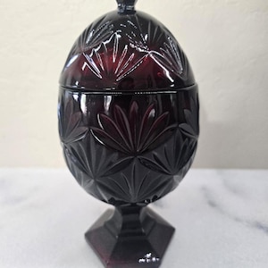 May include: A ruby-red, vintage glass candy dish with a lid. The dish features a detailed, cut-glass pattern of radiating leaves and a pedestal base. The lidded container is a decorative piece.