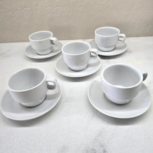 May include: A set of six white ceramic teacups and saucers. The cups have a simple, rounded design with a small handle. The saucers are round and slightly concave. The set is arranged on a light-colored surface.