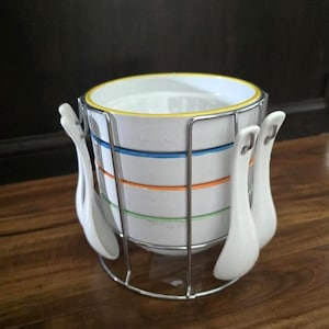 May include: A white ceramic soup bowl set with a metal rack and matching spoons. The bowl has blue, orange, and green stripes. The rim is yellow. The set is on a wooden surface.