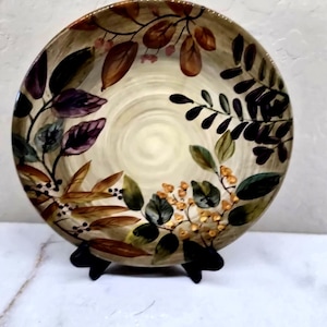 May include: A round ceramic plate featuring a cream-colored center with a spiral texture. The rim is decorated with painted botanical motifs, including dark purple, brown, and green leaves, along with clusters of small yellow and black berries.