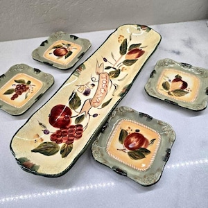 May include: A long, rectangular serving tray and four small square dishes made of ceramic. Each piece features a cream-colored center with painted red apples, grapes, and green leaves, surrounded by a sage green border. The tray displays the word TOSCANA.