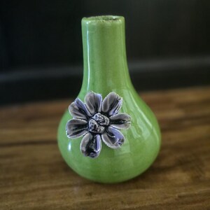 May include: A small, green ceramic vase with a glossy finish. A decorative, three-dimensional purple flower is affixed to the front. The vase has a rounded body and a narrow neck. The vase is sitting on a wooden surface.