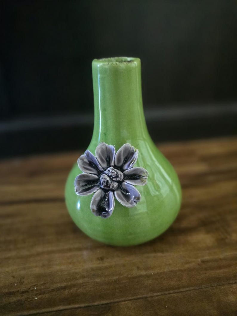 May include: A small, green ceramic vase with a glossy finish. A decorative, three-dimensional purple flower is affixed to the front. The vase has a rounded body and a narrow neck. The vase is sitting on a wooden surface.
