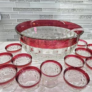 May include: A clear glass punch bowl set with a ruby red rim and matching glasses. The set includes a large bowl, a ladle, and several small glasses, perfect for serving beverages at a party or gathering.