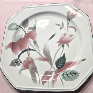 May include: An octagonal white plate with a floral design featuring pink roses and green leaves. The plate has a black trim and is set against a pink background. A decorative piece for dining.