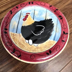 May include: A decorative ceramic plate featuring a hand-painted hen design. The plate has a red border with black leaf accents. The hen is black with a yellow head and red comb, and is surrounded by a blue and tan background.