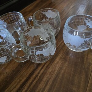 May include: Five clear glass mugs with handles, each featuring a world map design. The mugs are arranged on a wooden surface, showcasing their transparent quality and the detailed map etchings. The mugs are ideal for coffee or tea.