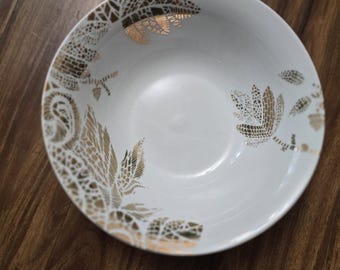 May include: A white ceramic bowl with a gold leaf pattern around the rim. The bowl has a smooth, glossy finish and is set against a dark wood surface. The gold design adds a touch of elegance.