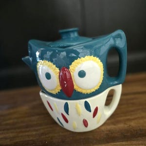 May include: A teal ceramic teapot and teacup set shaped like an owl. The teapot has a lid and handle, with yellow, white, and red accents. The teacup is white with colorful teardrop designs.