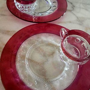 May include: Two clear glass snack sets, each consisting of a plate and a matching cup. The plates and cups feature a deep red rim and a textured, embossed pattern on the clear glass sections. The cups have a small handle and a circular design.