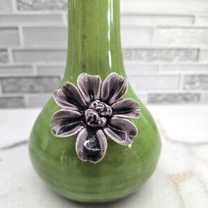May include: A green ceramic vase with a bulbous base and a slender neck. A detailed, dark purple flower is affixed to the front of the vase. The vase has a glossy finish and is suitable for displaying flowers or as a decorative item.