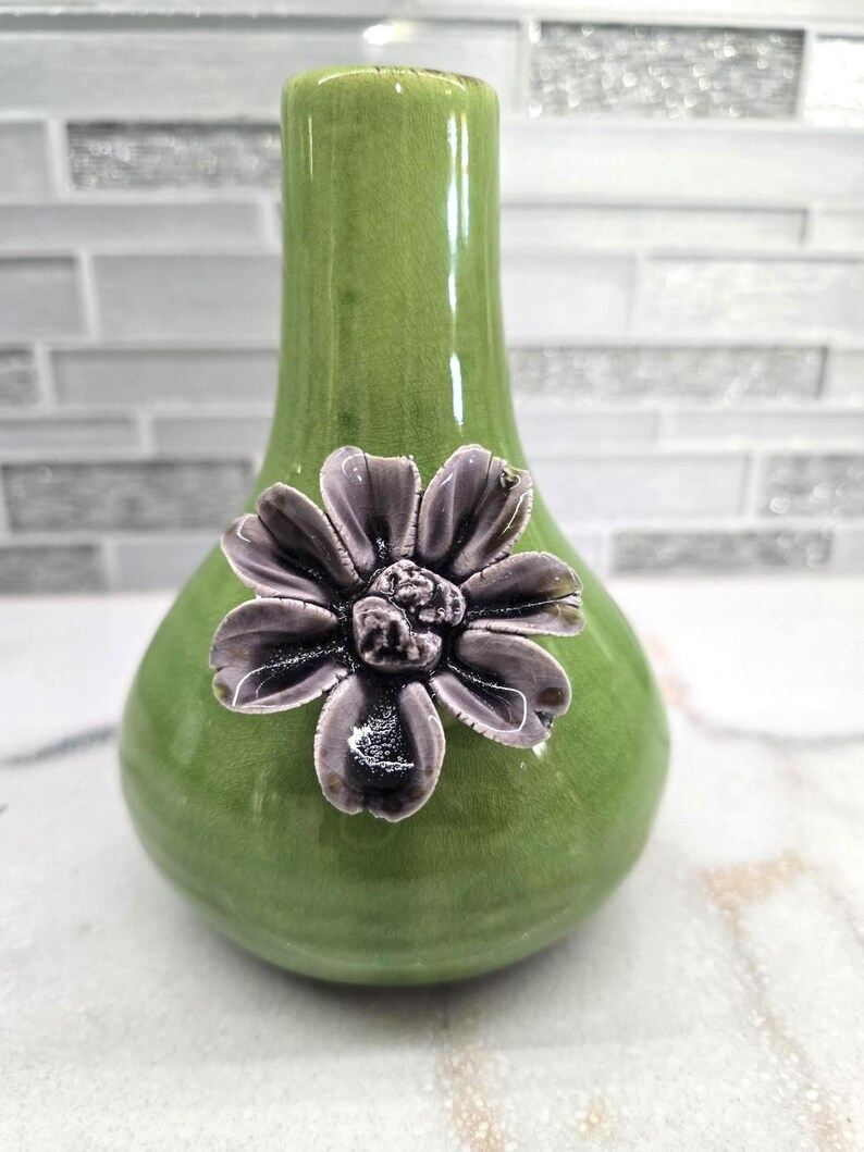May include: A green ceramic vase with a bulbous base and a slender neck. A detailed, dark purple flower is affixed to the front of the vase. The vase has a glossy finish and is suitable for displaying flowers or as a decorative item.