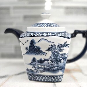 May include: A white and blue porcelain teapot with a landscape design. The teapot features a square body, a curved handle, and a lid. The landscape design includes mountains, trees, and buildings. The teapot is approximately 6 inches tall.