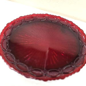 May include: A round, deep red glass plate featuring a textured, scalloped edge with an embossed chain-link pattern. The center of the plate displays a radiating sunburst design that extends outward from the middle toward the decorative rim.