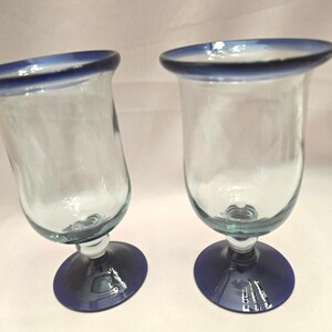 May include: Two clear glass goblets with a blue rim and base. The goblets have a rounded bowl shape and a short stem. The base is a solid, dark blue color. The rim of the glass is also blue.