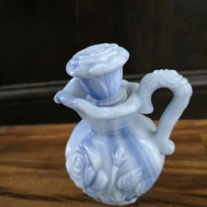 May include: A small, light blue and white glass pitcher with a rose-shaped stopper. The pitcher features a handle and is decorated with embossed roses and leaves. The design is a classic, vintage style.