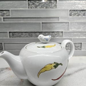 May include: A white ceramic teapot with a lid featuring a small bird figurine. The teapot is decorated with yellow chili pepper designs and a red curved line. The teapot has a spout and a handle.